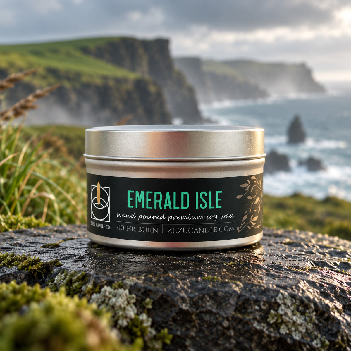 A silver metal tin candle labeled 'EMERALD ISLE' by Zuzuc Candle Co. sits on a mossy rock with a dramatic coastal cliff and ocean background.
